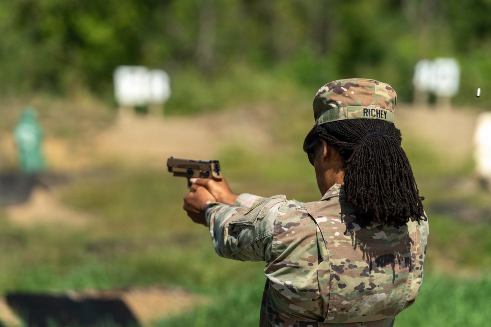 A U.S. Army soldier identified by name tape as 'RICHEY' is firing a pistol at a military shooting range. The soldier is wearing Operational Camouflage Pattern (OCP) uniform and patrol cap, and is using a two-handed grip stance. A spent casing is visible in the air to the right of the weapon, indicating the pistol has just been fired.