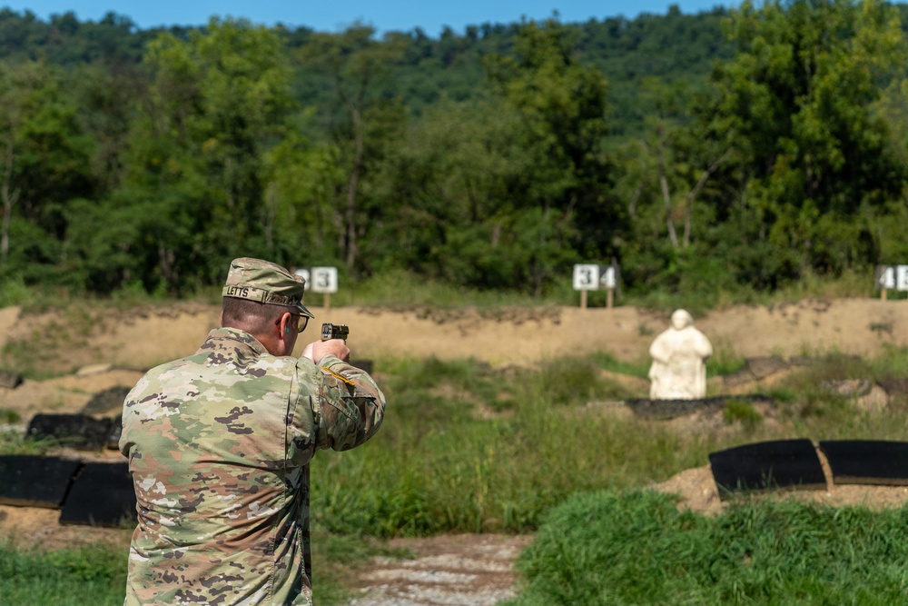 A soldier in U.S. Army OCP (Operational Camouflage Pattern) uniform is firing a pistol at an outdoor shooting range. The shooter is aiming at a white silhouette target positioned downrange, with numbered lane markers visible in the background. The setting appears to be a military qualification or training range during daytime.