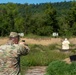 A soldier in U.S. Army OCP (Operational Camouflage Pattern) uniform is firing a pistol at an outdoor shooting range. The shooter is aiming at a white silhouette target positioned downrange, with numbered lane markers visible in the background. The setting appears to be a military qualification or training range during daytime.