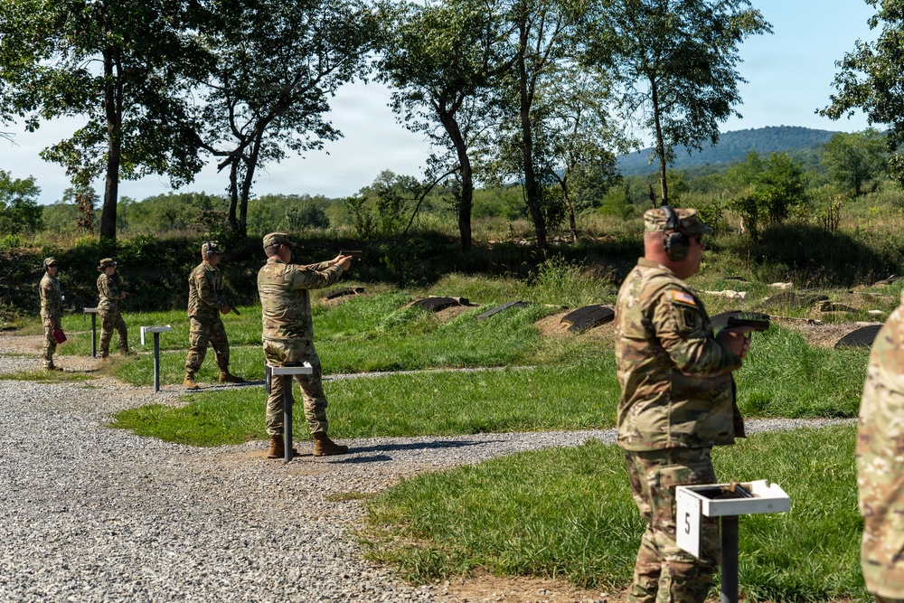 Military personnel in camouflage uniforms are conducting a pistol qualification or training exercise at an outdoor shooting range. Multiple soldiers are positioned at numbered firing lanes, with at least one actively aiming a pistol downrange. The setting appears to be a well-maintained military range with gravel pathways, numbered lane markers, and a natural wooded backdrop.
