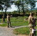 Military personnel in camouflage uniforms are conducting a pistol qualification or training exercise at an outdoor shooting range. Multiple soldiers are positioned at numbered firing lanes, with at least one actively aiming a pistol downrange. The setting appears to be a well-maintained military range with gravel pathways, numbered lane markers, and a natural wooded backdrop.