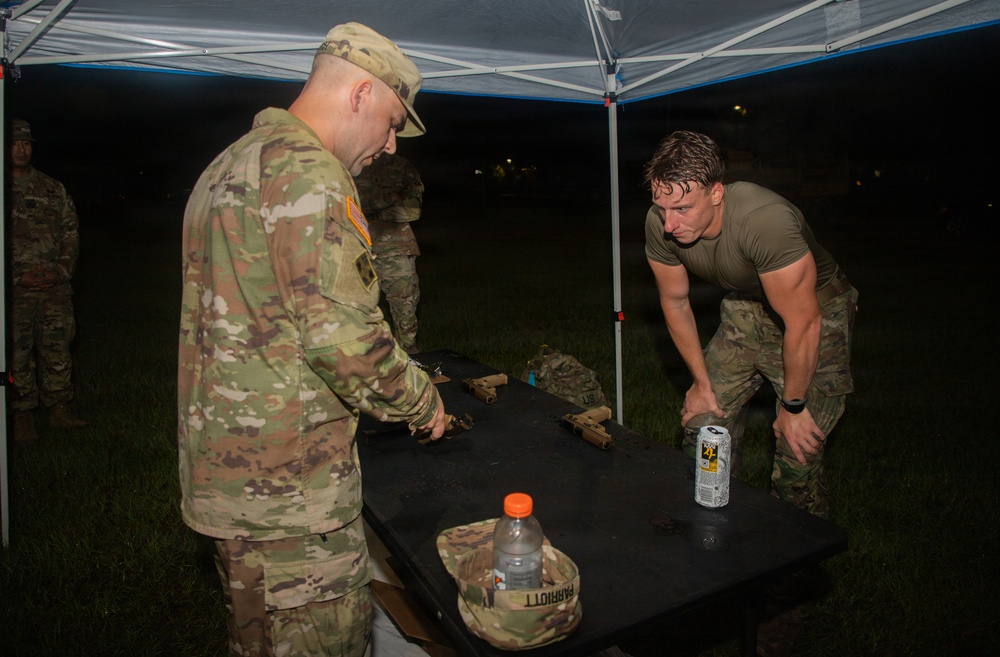Two U.S. Army soldiers are engaged in what appears to be a weapons inspection or equipment evaluation at a field station under a pop-up canopy tent at night. One soldier in full OCP (Operational Camouflage Pattern) uniform examines items on a black table, while a second soldier in a PT-style green shirt and OCP pants leans over the table. A name tape reading 'PARRIOTT' is visible on a patrol cap placed on the table.
