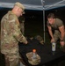 Two U.S. Army soldiers are engaged in what appears to be a weapons inspection or equipment evaluation at a field station under a pop-up canopy tent at night. One soldier in full OCP (Operational Camouflage Pattern) uniform examines items on a black table, while a second soldier in a PT-style green shirt and OCP pants leans over the table. A name tape reading 'PARRIOTT' is visible on a patrol cap placed on the table.