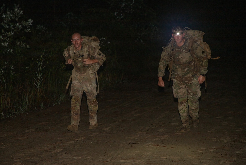 Two military personnel in camouflage uniforms are conducting a nighttime ruck march along a dirt trail. Both soldiers are carrying large rucksacks and appear to be moving at a fast pace, with one soldier wearing an illuminated headlamp. The scene is set in a dark, wooded environment lit primarily by the headlamp and camera flash.