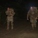 Two military personnel in camouflage uniforms are conducting a nighttime ruck march along a dirt trail. Both soldiers are carrying large rucksacks and appear to be moving at a fast pace, with one soldier wearing an illuminated headlamp. The scene is set in a dark, wooded environment lit primarily by the headlamp and camera flash.