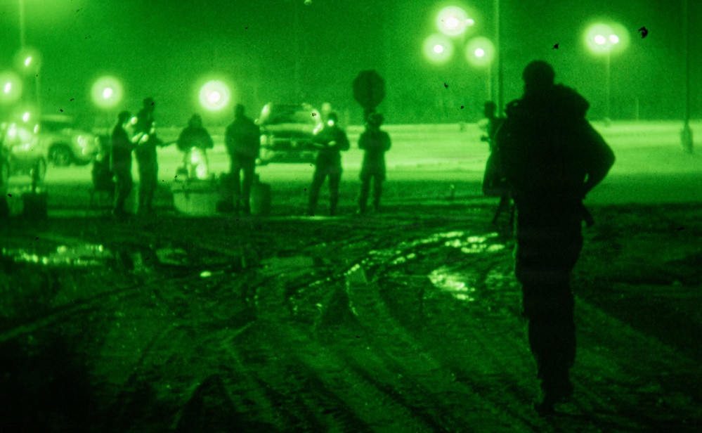 A night vision (NVG) image captured in green monochrome showing multiple individuals in an outdoor area, likely a parking lot or open ground. One armed individual is prominently silhouetted in the foreground, while a group of people stands in the middle distance near vehicles. The scene appears to depict a military or law enforcement operation conducted at night.