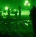A night vision (NVG) image captured in green monochrome showing multiple individuals in an outdoor area, likely a parking lot or open ground. One armed individual is prominently silhouetted in the foreground, while a group of people stands in the middle distance near vehicles. The scene appears to depict a military or law enforcement operation conducted at night.