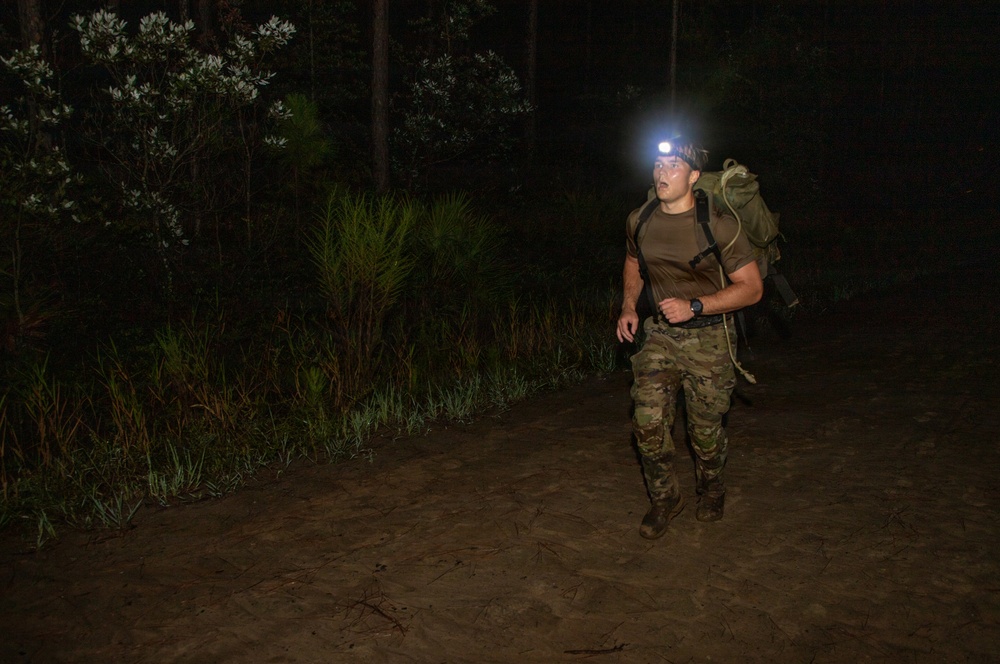 A male soldier or military trainee is conducting a nighttime ruck march or physical training exercise through a wooded, sandy terrain. He is wearing camouflage pants, a brown t-shirt, and carrying a large military rucksack while using a headlamp for illumination. The image was taken at night with the subject lit primarily by the headlamp and likely a camera flash.