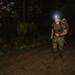 A male soldier or military trainee is conducting a nighttime ruck march or physical training exercise through a wooded, sandy terrain. He is wearing camouflage pants, a brown t-shirt, and carrying a large military rucksack while using a headlamp for illumination. The image was taken at night with the subject lit primarily by the headlamp and likely a camera flash.