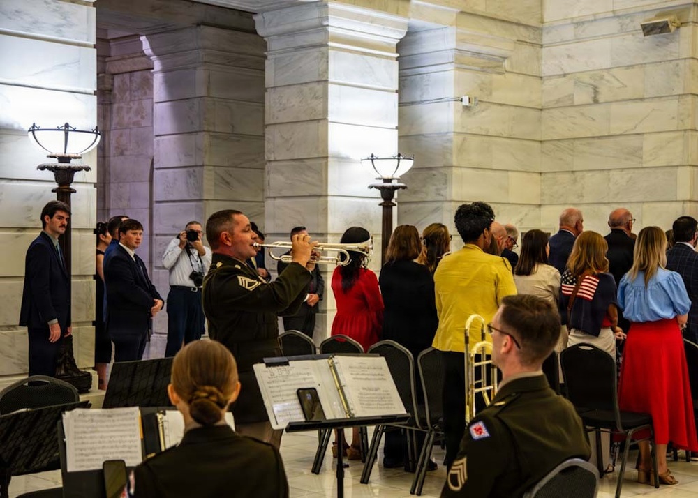 A formal military ceremony or official event is taking place inside a marble-columned government building, likely a state capitol rotunda. Military band members in Army dress uniforms are performing, with a trumpeter prominently playing while other musicians read sheet music. A crowd of formally dressed civilians and officials stands in attendance, with an American flag visible among the gathering.