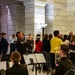 A formal military ceremony or official event is taking place inside a marble-columned government building, likely a state capitol rotunda. Military band members in Army dress uniforms are performing, with a trumpeter prominently playing while other musicians read sheet music. A crowd of formally dressed civilians and officials stands in attendance, with an American flag visible among the gathering.
