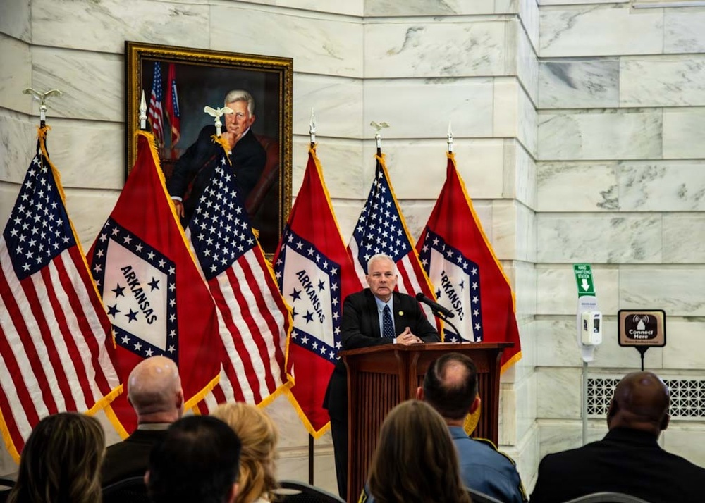A man in a dark suit speaks at a wooden podium during an official government event, likely at the Arkansas State Capitol based on the marble interior and prominent Arkansas state flags. Multiple alternating Arkansas state flags and American flags are displayed behind the speaker, with a formal oil portrait of an unidentified official hanging on the wall above. An audience is seated in the foreground with their backs to the camera.