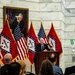 A man in a dark suit speaks at a wooden podium during an official government event, likely at the Arkansas State Capitol based on the marble interior and prominent Arkansas state flags. Multiple alternating Arkansas state flags and American flags are displayed behind the speaker, with a formal oil portrait of an unidentified official hanging on the wall above. An audience is seated in the foreground with their backs to the camera.
