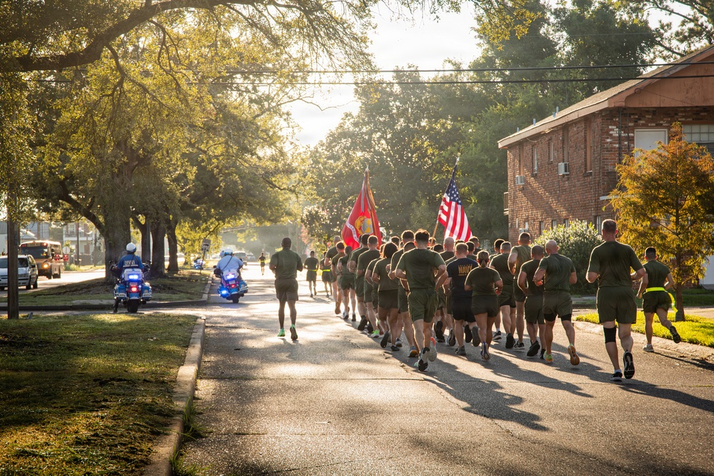 A large formation of military personnel, appearing to be U.S. Marines based on their olive drab PT (physical training) uniforms, are conducting a group run along a tree-lined residential street. Two flag bearers carry the U.S. Marine Corps flag and the American flag at the front of the formation. The run is escorted by police motorcycles with blue lights activated, suggesting an official or ceremonial event.