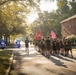 A large formation of military personnel, appearing to be U.S. Marines based on their olive drab PT (physical training) uniforms, are conducting a group run along a tree-lined residential street. Two flag bearers carry the U.S. Marine Corps flag and the American flag at the front of the formation. The run is escorted by police motorcycles with blue lights activated, suggesting an official or ceremonial event.