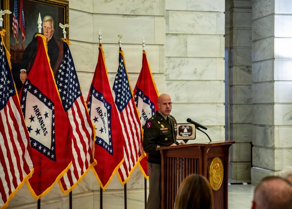 A U.S. Army officer in green service uniform is speaking at a wooden podium inside what appears to be the Arkansas State Capitol rotunda. Multiple Arkansas state flags and U.S. flags are displayed behind him, alongside a formal portrait painting of a dignitary on the wall. The setting suggests an official military or governmental ceremony.