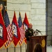 A U.S. Army officer in green service uniform is speaking at a wooden podium inside what appears to be the Arkansas State Capitol rotunda. Multiple Arkansas state flags and U.S. flags are displayed behind him, alongside a formal portrait painting of a dignitary on the wall. The setting suggests an official military or governmental ceremony.