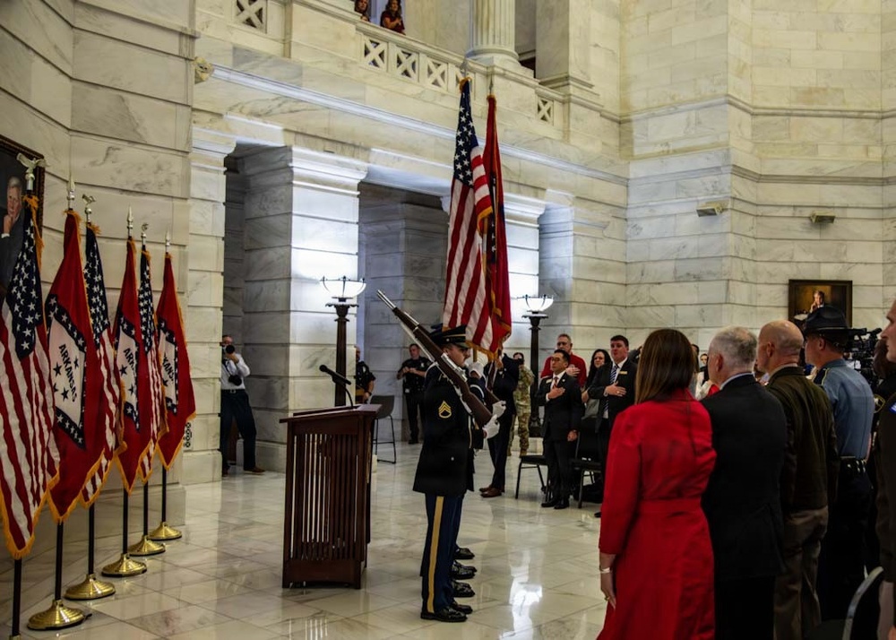 A formal military color guard ceremony is taking place inside what appears to be a state capitol rotunda, identifiable by its marble columns and classical architecture. A soldier in dress uniform presents the U.S. flag and what appears to be the Arkansas state flag while attendees stand at attention with hands over hearts. Multiple Arkansas state flags and U.S. flags are displayed on stands to the left, suggesting an official government ceremony such as an inauguration or swearing-in.