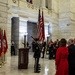 A formal military color guard ceremony is taking place inside what appears to be a state capitol rotunda, identifiable by its marble columns and classical architecture. A soldier in dress uniform presents the U.S. flag and what appears to be the Arkansas state flag while attendees stand at attention with hands over hearts. Multiple Arkansas state flags and U.S. flags are displayed on stands to the left, suggesting an official government ceremony such as an inauguration or swearing-in.