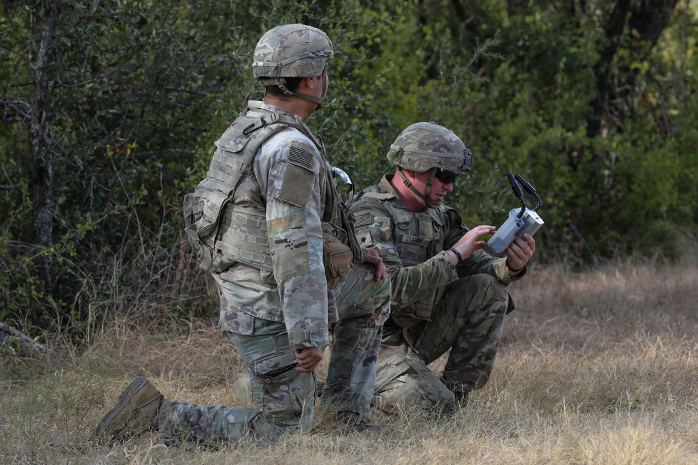 Two military personnel in Operational Camouflage Pattern (OCP) uniforms are positioned in a field near treeline vegetation. One soldier is kneeling and operating what appears to be a small drone or electronic device, while the other stands nearby observing. The setting suggests a field training exercise or operational deployment.