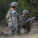 Two military personnel in Operational Camouflage Pattern (OCP) uniforms are positioned in a field near treeline vegetation. One soldier is kneeling and operating what appears to be a small drone or electronic device, while the other stands nearby observing. The setting suggests a field training exercise or operational deployment.