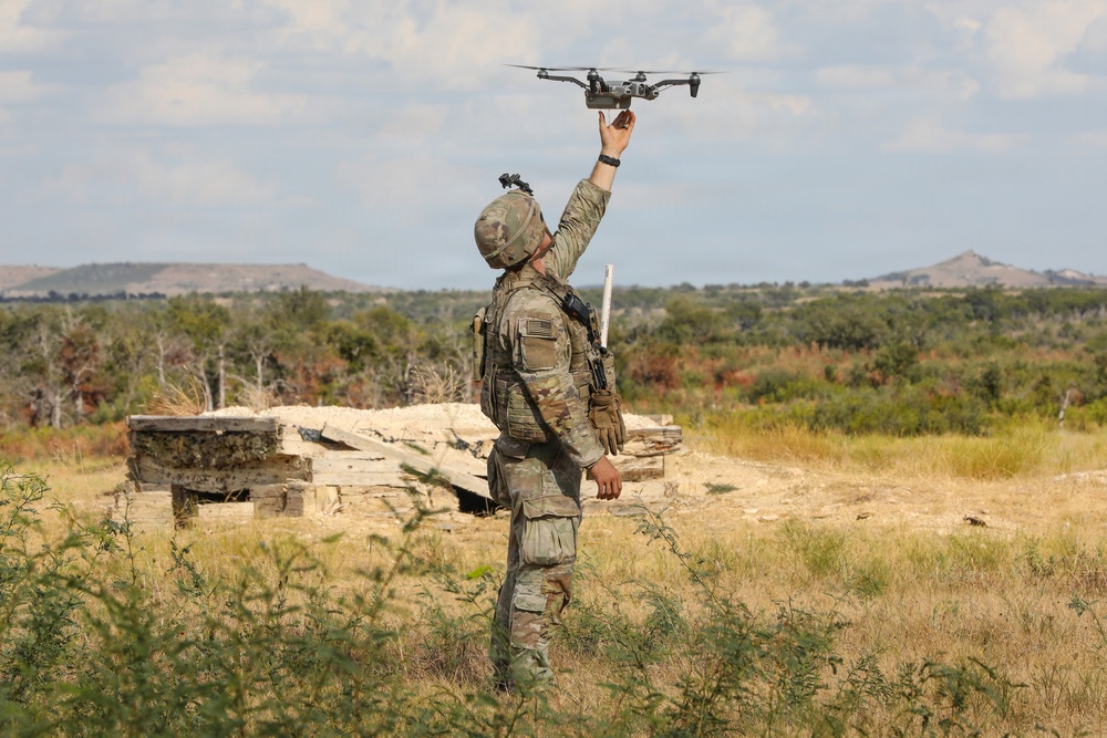 A soldier in full camouflage combat gear is launching or recovering a small quadcopter drone in an arid, scrubland training environment. The individual is holding the drone aloft with one raised arm against a backdrop of low vegetation, distant hills, and a partially collapsed wooden structure. The scene appears consistent with a military field training exercise involving unmanned aerial systems (UAS).
