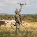A soldier in full camouflage combat gear is launching or recovering a small quadcopter drone in an arid, scrubland training environment. The individual is holding the drone aloft with one raised arm against a backdrop of low vegetation, distant hills, and a partially collapsed wooden structure. The scene appears consistent with a military field training exercise involving unmanned aerial systems (UAS).