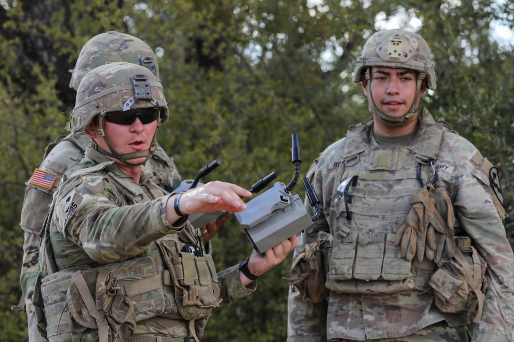 Two U.S. Army soldiers in full combat gear are engaged in what appears to be a training exercise involving a small drone or UAS (Unmanned Aerial System) controller. The soldier on the left, wearing sunglasses and identified by a name tape reading 'JACOBS,' is holding and explaining a compact drone device to the soldier on the right. Both are wearing OCP (Operational Camouflage Pattern) uniforms with tactical vests and helmets.