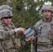 Two U.S. Army soldiers in full combat gear are engaged in what appears to be a training exercise involving a small drone or UAS (Unmanned Aerial System) controller. The soldier on the left, wearing sunglasses and identified by a name tape reading 'JACOBS,' is holding and explaining a compact drone device to the soldier on the right. Both are wearing OCP (Operational Camouflage Pattern) uniforms with tactical vests and helmets.