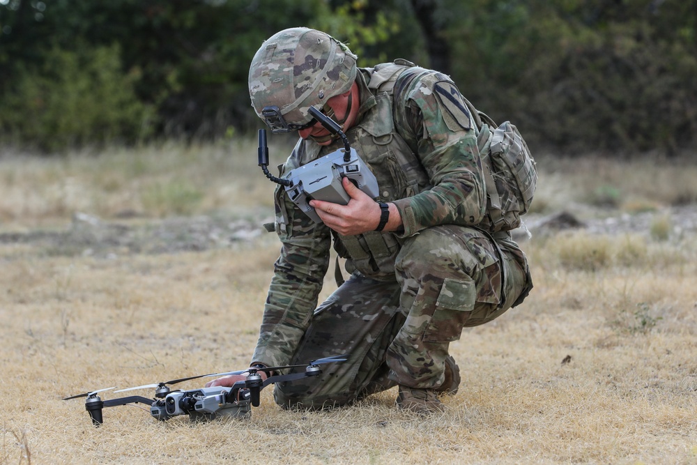 A military soldier in full camouflage (OCP/MultiCam) uniform is crouched on dry grassland preparing or operating a small quadcopter drone. The soldier is holding a remote control unit and appears to be configuring or launching the unmanned aerial vehicle (UAV). The setting appears to be a field training exercise based on the terrain and equipment.