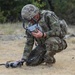 A military soldier in full camouflage (OCP/MultiCam) uniform is crouched on dry grassland preparing or operating a small quadcopter drone. The soldier is holding a remote control unit and appears to be configuring or launching the unmanned aerial vehicle (UAV). The setting appears to be a field training exercise based on the terrain and equipment.