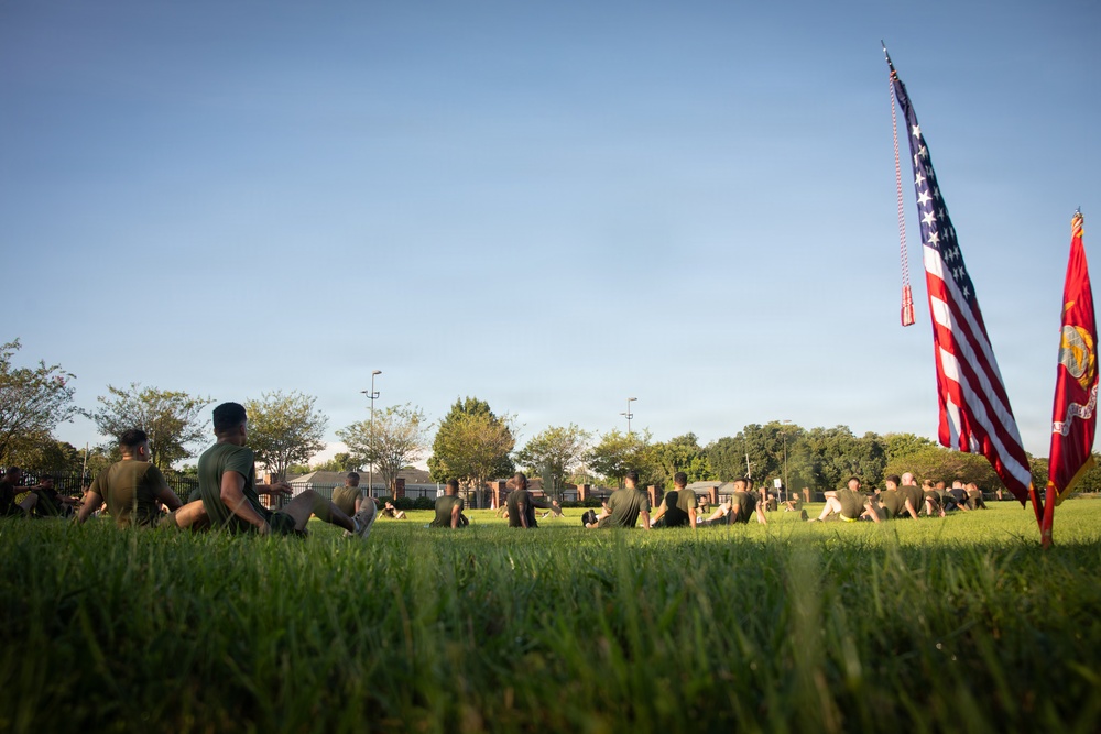 A group of military personnel in olive drab physical training uniforms are performing exercises on a grass field, likely during a morning physical training session. Two flags are prominently displayed on the right side: the United States flag and what appears to be a United States Marine Corps flag. The photo is taken from a low ground-level angle during what appears to be early morning based on the warm directional lighting.