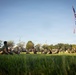 A group of military personnel in olive drab physical training uniforms are performing exercises on a grass field, likely during a morning physical training session. Two flags are prominently displayed on the right side: the United States flag and what appears to be a United States Marine Corps flag. The photo is taken from a low ground-level angle during what appears to be early morning based on the warm directional lighting.