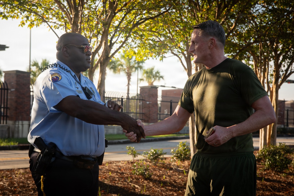 Two men are shaking hands outdoors near what appears to be a secured facility entrance with brick pillars and iron fencing. One man is wearing a light blue law enforcement or security uniform with a badge and duty belt, while the other is dressed in olive/dark green military-style physical training attire. The interaction appears cordial and formal, suggesting a professional greeting or acknowledgment.
