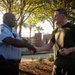 Two men are shaking hands outdoors near what appears to be a secured facility entrance with brick pillars and iron fencing. One man is wearing a light blue law enforcement or security uniform with a badge and duty belt, while the other is dressed in olive/dark green military-style physical training attire. The interaction appears cordial and formal, suggesting a professional greeting or acknowledgment.