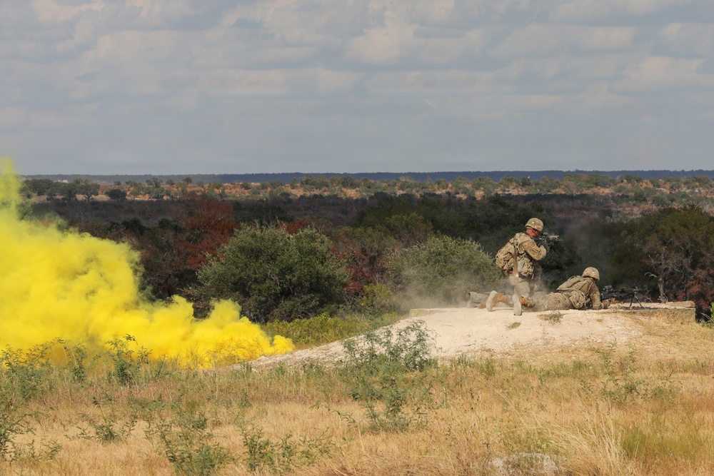 Military personnel are conducting a field training exercise on elevated rocky terrain, with a yellow smoke grenade deployed to the left, likely used for signaling or marking purposes. Two soldiers in desert camouflage uniforms are visible — one standing and moving, one in a prone position — suggesting a tactical drill. The landscape is arid scrubland consistent with a training range in the American Southwest.