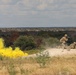 Military personnel are conducting a field training exercise on elevated rocky terrain, with a yellow smoke grenade deployed to the left, likely used for signaling or marking purposes. Two soldiers in desert camouflage uniforms are visible — one standing and moving, one in a prone position — suggesting a tactical drill. The landscape is arid scrubland consistent with a training range in the American Southwest.