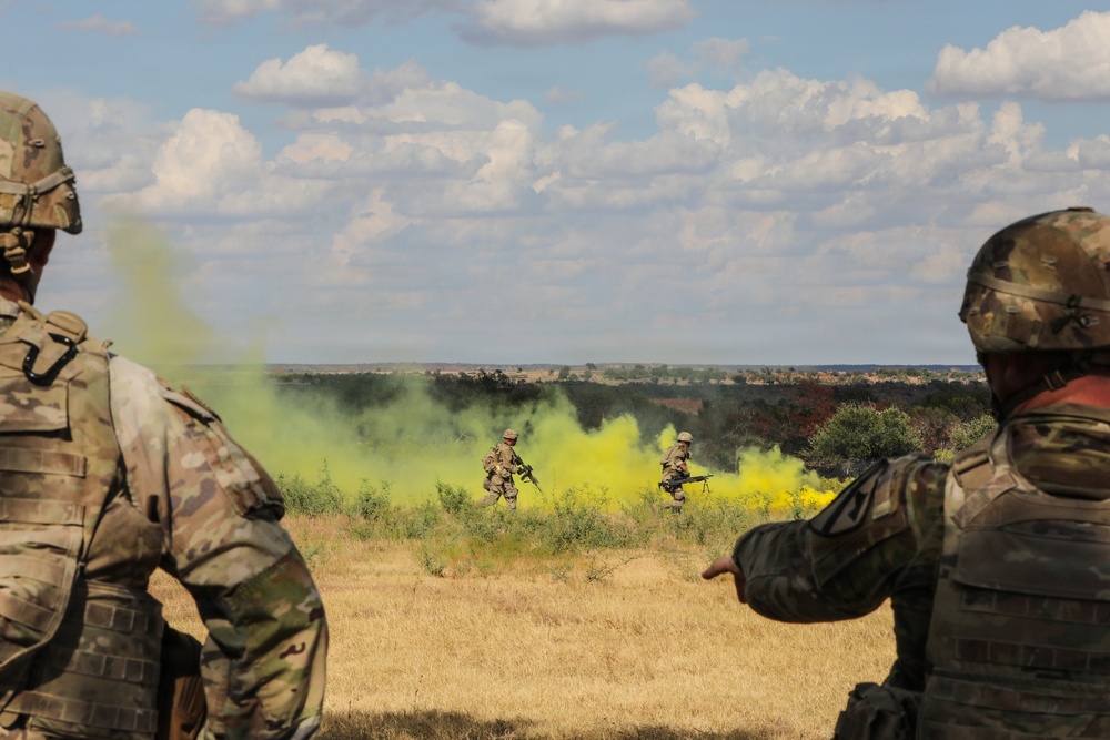 Military personnel are conducting a field training exercise in an open, arid landscape. Yellow smoke grenades have been deployed, likely for signaling or marking purposes during the drill. Two soldiers in the foreground observe while two others maneuver through the smoke in the middle ground.