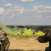 Military personnel are conducting a field training exercise in an open, arid landscape. Yellow smoke grenades have been deployed, likely for signaling or marking purposes during the drill. Two soldiers in the foreground observe while two others maneuver through the smoke in the middle ground.