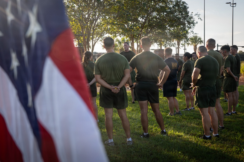 A group of military personnel in physical training (PT) uniforms are gathered outdoors on a grass field, appearing to receive a briefing or instruction from a leader standing in the center. An American flag is prominently visible in the foreground left, partially out of focus. The setting appears to be an early morning formation or pre-workout assembly at a military installation.