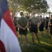 A group of military personnel in physical training (PT) uniforms are gathered outdoors on a grass field, appearing to receive a briefing or instruction from a leader standing in the center. An American flag is prominently visible in the foreground left, partially out of focus. The setting appears to be an early morning formation or pre-workout assembly at a military installation.