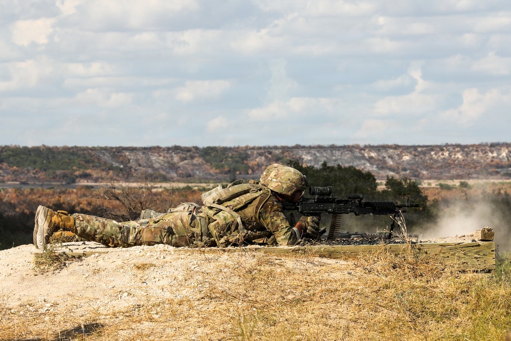 A soldier in full camouflage uniform and combat gear is in a prone firing position on an elevated dirt berm, operating what appears to be an M249 or M240 light machine gun. Smoke or dust is visible near the muzzle area, suggesting the weapon has recently been or is being fired. The setting appears to be a military training range in an arid, scrubland environment.