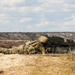 A soldier in full camouflage uniform and combat gear is in a prone firing position on an elevated dirt berm, operating what appears to be an M249 or M240 light machine gun. Smoke or dust is visible near the muzzle area, suggesting the weapon has recently been or is being fired. The setting appears to be a military training range in an arid, scrubland environment.