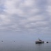 A large naval warship, consistent with a US Navy Littoral Combat Ship (trimaran/Independence-class design), is underway on calm, overcast coastal waters. A smaller escort or patrol boat accompanies it on its starboard side, leaving a visible wake. Several small fishing or civilian vessels are scattered in the background to the left.