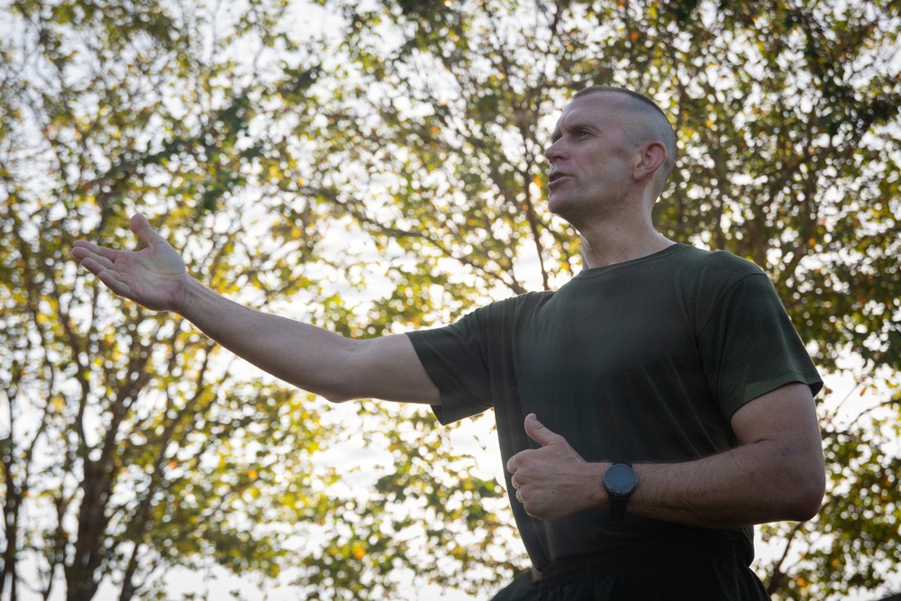 A middle-aged man with a close-cropped hairstyle is photographed outdoors from a low angle against a backdrop of trees with backlit foliage. He is wearing a dark olive/military green t-shirt and a black wristwatch, with one arm extended outward in a gesturing or instructional pose. The lighting suggests early morning or late afternoon sun filtering through the trees.