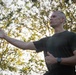 A middle-aged man with a close-cropped hairstyle is photographed outdoors from a low angle against a backdrop of trees with backlit foliage. He is wearing a dark olive/military green t-shirt and a black wristwatch, with one arm extended outward in a gesturing or instructional pose. The lighting suggests early morning or late afternoon sun filtering through the trees.