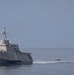 A trimaran-hulled Littoral Combat Ship (LCS) is anchored or underway at slow speed on calm open water under a hazy sky. The vessel displays a red and white flag on its mast and features the distinctive angular, low-profile superstructure characteristic of the Independence-class LCS. Two large seabirds, likely pelicans, are visible in flight in the foreground to the right of the ship.