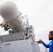 A man wearing a blue long-sleeve shirt is interacting with a large gray military weapons system, likely a Phalanx Close-In Weapon System (CIWS) or similar naval defense turret. The equipment features a distinctive dome-shaped radar housing mounted on a boxy gray pedestal structure. The setting appears to be on or near a naval vessel or military installation under an overcast sky.