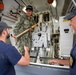 A U.S. Navy sailor in digital camouflage uniform holds a large brass artillery shell/round inside what appears to be a naval gun mount or weapons system compartment. Two civilians wearing shirts with 'ISEA' (In-Service Engineering Activity) logos observe or interact with the sailor. The scene appears to be a demonstration, training, or inspection activity aboard a naval vessel.