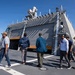 A group of civilians are touring the deck of a modern naval warship, likely during a public open-ship event or official visit. The vessel features a distinctive angular, stealth-design superstructure with radar systems and weapons mounts visible on top. The setting appears to be a naval port or shipyard on a clear, sunny day.
